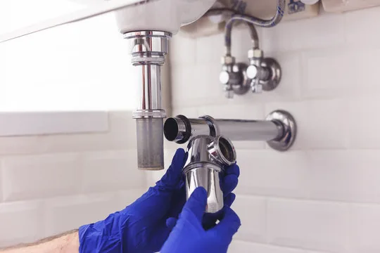Plumber in blue gloves removing a chrome sink P-trap to clear a clog