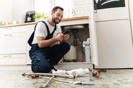 Smiling plumber kneeling at an open kitchen sink cabinet, checking his phone beside pipes and parts